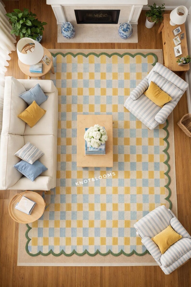 May include: An aerial view of a living room featuring a large rug with a yellow and pale blue chequered pattern and a green scalloped border. A white sofa, striped armchairs, and wooden tables are arranged around the rug. Cushions in blue and yellow.