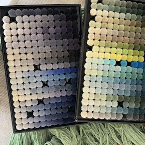 May include: Two black trays displaying small, round carpet samples in a range of colours. One tray shows a gradient of blues and greys, the other greens, yellows, and blues. Green and orange yarn are visible below the trays.