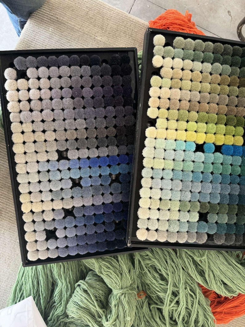 May include: Two black trays displaying small, round carpet samples in a range of colours. One tray shows a gradient of blues and greys, the other greens, yellows, and blues. Green and orange yarn are visible below the trays.