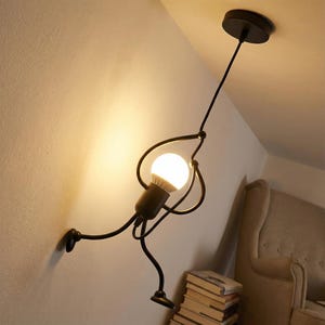 Pode incluir: Uma luminária pendente de metal preto peculiar, projetada como uma figura de palito abraçando uma lâmpada. A luz é montada em uma parede branca e suspensa do teto por um cabo preto. A lâmpada está acesa.