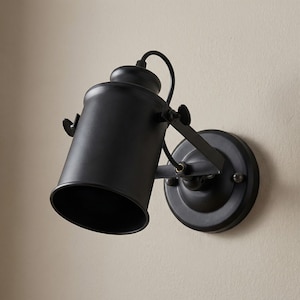 May include: A black, industrial-style wall-mounted spotlight. The lamp features a cylindrical shade, adjustable arm, and a circular base. The fixture is made of metal and has a matte finish. The spotlight is designed to direct light.