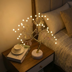 Puede incluir: Lámpara decorativa en forma de árbol con luces LED blancas cálidas. Las ramas de metal dorado presentan detalles en forma de hojas. La lámpara está sobre una base blanca, en una mesita de noche de madera.