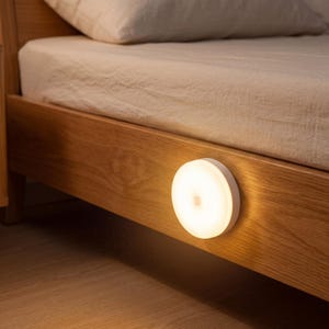 May include: A round, white LED night light is mounted on the side of a wooden bed frame. The light is illuminated, casting a warm glow on the wood and floor. The bed has a beige sheet and pillow.