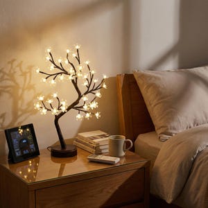 May include: A tree-shaped lamp with white flower lights. The lamp has a dark brown base and branches, and sits on a wooden bedside table. A digital weather station, books, a mug, and a remote control are also on the table. The bed is nearby.