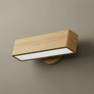 May include: A rectangular wooden wall sconce with a light-colored wood grain finish. The sconce has a recessed, rectangular light source with a white glow. The fixture is mounted on a circular wooden base.