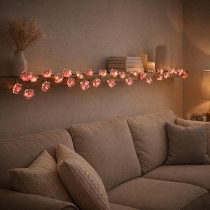 Può includere: Una stringa di luci a forma di fiori rosa illumina una mensola di legno, creando un'atmosfera calda. Le luci sono sopra un divano beige. Un vaso con fiori secchi e libri sono anche sulla mensola.