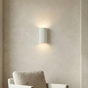May include: A white, cylindrical wall sconce with vertical ribbed detailing. The light fixture is mounted on a neutral-toned wall, emitting a warm glow. The sconce is a modern lighting fixture.