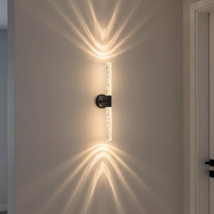 May include: A modern wall sconce with a black base and a vertical, clear, textured light fixture. The sconce emits light upwards and downwards, creating a starburst effect on the wall. The fixture is mounted on a neutral-colored wall.