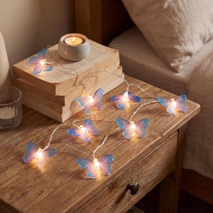 Pode incluir: Uma sequência de luzes em forma de borboleta azul e roxa, iluminadas com luz branca quente. As luzes estão dispostas numa mesa de cabeceira de madeira, ao lado de livros e uma vela acesa.