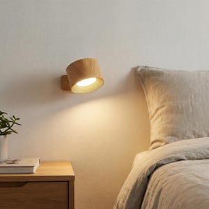 May include: A wooden wall-mounted lamp with a cylindrical shade and a warm, inviting glow. The lamp is made of light-colored wood and is mounted on a matching wooden base. The lamp is positioned on a white wall, next to a bed.
