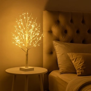 May include: A decorative LED birch tree lamp with warm white lights. The tree is on a small, round table next to a bed with a tufted headboard and pillows. The tree's trunk is wrapped in twine, and the base is square.
