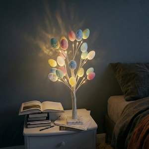 Op de afbeelding: Een decoratieve paaseierenboom met veelkleurige eieren en warmwitte LED-verlichting. De boom staat op een witte basis en staat op een nachtkastje. De eieren zijn pastelkleurig, waaronder roze, geel, blauw en groen.