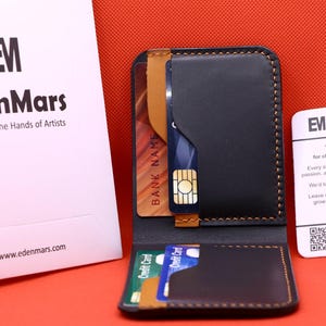 May include: Black and brown leather wallet with card slots, holding credit cards and a SIM card. The wallet is next to a white card with the EdenMars logo and a thank you message. A white bag with the EdenMars logo is also in the image.