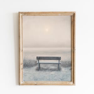 May include: A framed landscape print featuring a serene scene. A wooden bench sits in the foreground, facing a misty body of water under a pale sun. The artwork is framed in a rustic, light-toned wooden frame, creating a calming aesthetic.