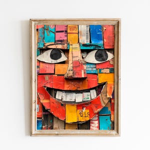 May include: A framed artwork featuring a smiling face constructed from colorful cardboard pieces. The face has large white eyes with black pupils, a prominent nose, and a wide red mouth. The artwork is framed in a light-colored wooden frame.