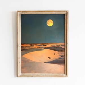 Puede incluir: Pintura de paisaje enmarcada que representa una escena desértica bajo un cielo nocturno. Una luna llena y amarilla ilumina las dunas. La pintura está en un marco de madera clara y envejecida.