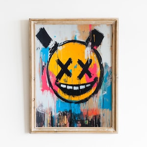 May include: Abstract artwork featuring a yellow smiley face with black cross-eyes and a wide grin. The painting has black, pink, blue, and orange paint splatters. The artwork is framed in a light brown wooden frame.