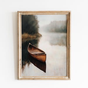 May include: A framed art print depicts a wooden canoe floating on a calm lake. The artwork features a muted color palette with a misty atmosphere, creating a serene and natural scene. The frame is a light wood color.
