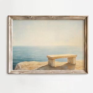 May include: A framed print showing a coastal scene. A stone bench is on a cliff overlooking a calm, blue sea under a soft, yellow sky. The frame is made of light-coloured wood.