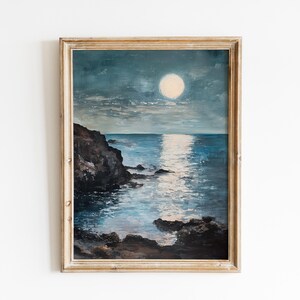 May include: A framed vintage painting depicts a moonlit seascape. The artwork features a large, bright moon reflecting on the water, with dark rocky cliffs in the foreground. The color palette includes blues, whites, and browns.