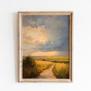 May include: A framed landscape painting depicts a path winding through a field of golden wheat under a dramatic sky. The artwork features a warm color palette with shades of yellow, orange, and blue, creating a serene and inviting scene. The frame is a light, distressed wood.