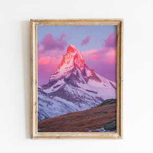 May include: Framed print of a snow-covered mountain peak at sunset. The mountain is lit with pink and orange hues, contrasting with the blue and purple sky. The frame is a light brown color, and the landscape includes a grassy hillside.