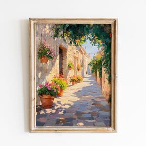 May include: Framed painting of a sunlit European alleyway. Stone buildings with flower pots and climbing plants line the cobbled street. The artwork features warm colours and a sense of depth, creating a picturesque scene.