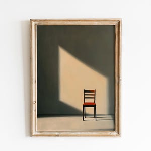 May include: A framed art print featuring a minimalist interior scene. The artwork depicts a single wooden chair in a room with a light-filled corner. The chair has a red seat and a dark brown frame. The frame around the artwork is a light brown color.