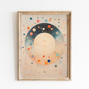 Op de afbeelding: Een ingelijste kunstdruk met een hemels thema. Het kunstwerk toont concentrische cirkels, met een centrale, lichte cirkel en een ring van blauwe, oranje en witte stippen tegen een donkerblauwe achtergrond. De lijst is lichtbruin.