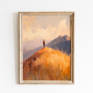 May include: Framed landscape art print featuring a person standing on a golden-hued hill, overlooking distant mountains. The artwork has a warm color palette with shades of orange, brown, and blue. The frame is a light, distressed wood.