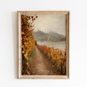 May include: Framed vintage landscape art print. A path winds through a vineyard with golden autumn leaves, leading towards a misty river and distant hills. The artwork is in a distressed, light-colored wooden frame.