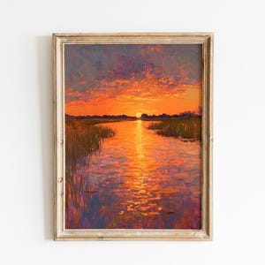 May include: Oil painting of a sunset over a river or marsh. The sky is a blend of orange, purple, and blue hues. The water reflects the sunset, with tall grasses along the banks. The painting is framed in a light-colored wooden frame.
