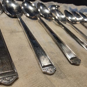 May include: A collection of vintage silver spoons with ornate, geometric designs on the handles. The spoons are arranged on a light-colored cloth, showcasing their reflective surfaces and classic style.