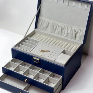 Könnte beinhalten: Eine marineblaue Schmuckschatulle mit mehreren Fächern und Schubladen. Der Innenraum ist mit einem hellgrauen Stoff ausgekleidet. Die Schatulle hat einen goldfarbenen Verschluss und einen kleinen schlüsselförmigen Anhänger. Die Schatulle ist geöffnet und zeigt ihre Stauräume.