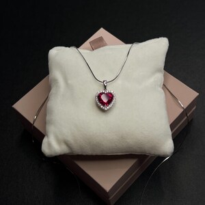 Op de afbeelding: Een zilveren ketting met een hartvormige hanger. De hanger heeft een grote, rode edelsteen omringd door kleinere, heldere stenen. De ketting wordt gepresenteerd op een wit kussen in een roze geschenkdoos, tegen een donkere achtergrond.