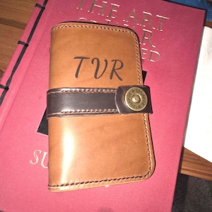 May include: A tan leather wallet featuring the letters TVR embossed on the front. The wallet is secured by a dark brown leather strap with a brass-colored metal snap button that displays the text 12 GAUGE.