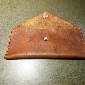 May include: A brown leather envelope-style wallet with a snap closure. The wallet is open, revealing the interior. The leather has a textured appearance and is stitched along the edges. A metal snap secures the flap.