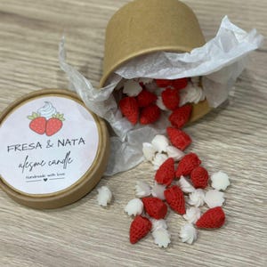 Puede incluir: Una vela con temática de fresa y nata. La vela está en un recipiente pequeño y redondo con tapa. La tapa tiene las palabras "FRESA & NATA" y "afesme candle" impresas. Alrededor hay fresas rojas y trozos de cera con forma de crema blanca.