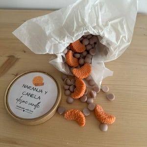 Puede incluir: Un recipiente redondo con el texto "NARANJA Y CANELA afesme candle" está junto a una bolsa de papel blanca. La bolsa derrama ceras perfumadas naranjas y marrones con forma de gajos de naranja y pequeños discos.