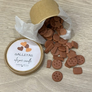 Puede incluir: Un recipiente de papel marrón con tapa, lleno de ceras perfumadas con forma de galleta marrón, derramándose sobre una superficie de madera. La tapa tiene la inscripción "GALLETAS afesme candle Handmade with love".