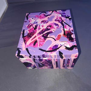 May include: A square wooden box with a colorful abstract design. The box is painted in shades of pink, purple, and black. The design is reminiscent of splattered paint.