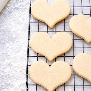 Puede incluir: Varias galletas en forma de corazón sobre una rejilla negra para enfriar. Las galletas son de color dorado claro y parecen recién horneadas. Harina ligeramente espolvoreada sobre la superficie gris. Un rodillo blanco en el fondo.
