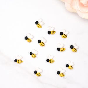 May include: A collection of small, decorative bee-shaped embellishments. Each bee features white wings, a black body, and yellow and black striped details. The bees are scattered on a white surface, with a soft pink rose in the background.