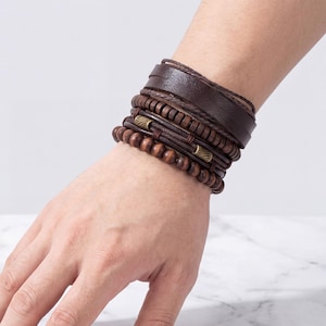May include: A collection of brown leather and wooden bead bracelets. The bracelets include a wide wrap-around leather cuff, braided leather bands, and beaded strands. The bracelets are stacked on a wrist, showcasing the different textures and styles.