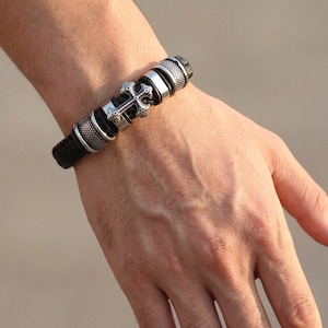 Op de afbeelding: Zwarte leren armband met een zilveren kruisbedel. De armband heeft een gevlochten leren band en zilveren accenten. Het kruis is gedetailleerd met ingewikkelde patronen. Een stijlvol accessoire.