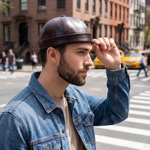 Puede incluir: Gorra de cuero marrón oscuro con una corona redondeada y un borde corto. La gorra la lleva una persona que lleva una chaqueta vaquera. El fondo muestra una calle de la ciudad con taxis amarillos y peatones.