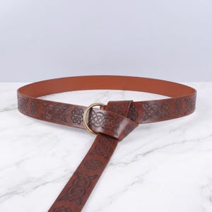 Puede incluir: Un cinturón de cuero marrón con intrincados nudos celtas en relieve. El cinturón presenta una hebilla circular de color latón y una larga correa decorativa. El cinturón está diseñado para usarse alrededor de la cintura.