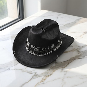 May include: A black cowboy hat featuring a textured surface, a black and white patterned band, and a silver-toned metal chain accent. The hat is adorned with several small silver metal rings attached to the crown and the wide, curved brim.