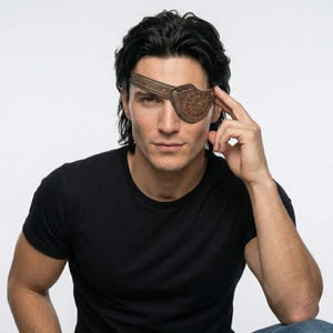 May include: A brown leather eye patch with a decorative design, worn over one eye. The eye patch is secured with a strap that goes around the head. The person in the image is wearing a black t-shirt.