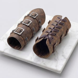 May include: Two brown leather arm guards with metal buckles and decorative studs. One arm guard has three buckles, while the other features a laced design. The arm guards are designed to protect the forearm.